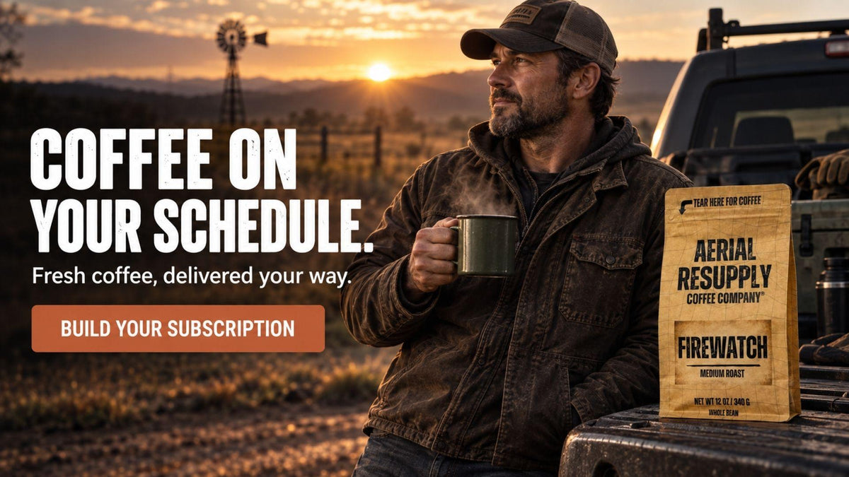 A man in a cap and jacket stands beside a truck at sunset holding a coffee mug, with large text reading 'COFFEE ON YOUR SCHEDULE.' and a wooden box labeled 'AERIAL RESUPPLY COFFEE COMPANY FIREWATCH' on the truck bed.