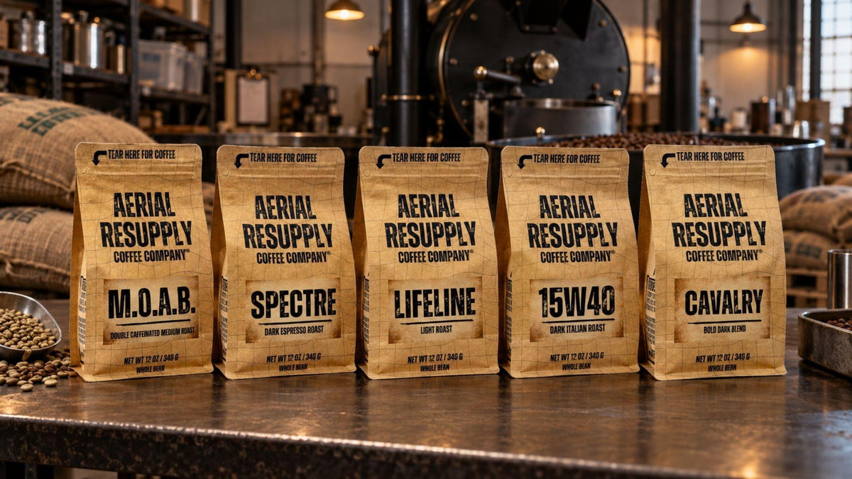 Five stand-up coffee bags from Aerial Resupply Coffee Company lined up on a countertop in a coffee shop, with burlap sacks and a coffee roaster blurred in the background.
