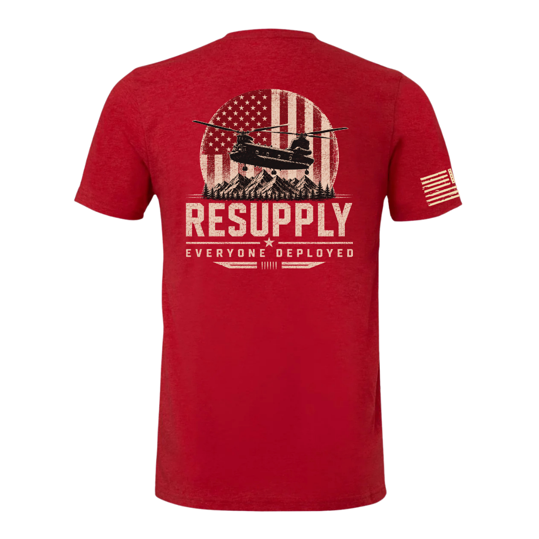 A red short-sleeve t-shirt shown from the back, featuring a distressed graphic of a military helicopter in front of an American flag and mountains, with the text 'RESUPPLY' and 'EVERYONE DEPLOYED'. A small American flag is printed on the left sleeve.